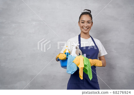 Young happy woman, female cleaner in uniform and rubber gloves holding bucket of cleaning supplies and smiling at camera, standing against grey wall Young happy woman, female cleaner in uniform and rubber gloves holding bucket of cleaning supplies and smiling at camera, standing against grey wall 68276924