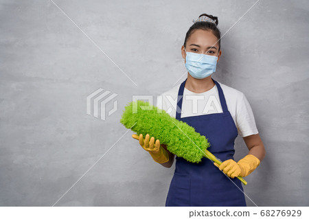 Confident cleaning lady wearing yellow rubber gloves and medical mask holding microfiber duster for cleaning and looking at camera, standing against grey wall Confident cleaning lady wearing yellow rubber gloves and medical mask holding microfiber duster for cleaning and looking at camera, standing against grey wall 68276929