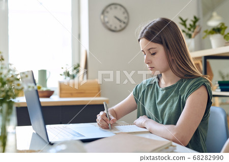 Serious teenage girl making notes while sitting by desk in front of laptop 68282990