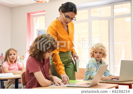 Elegant female teacher explaining work on laptop to children Elegant female teacher explaining work on laptop to children 68288254