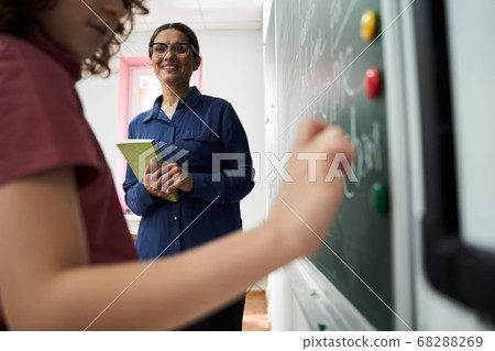 female teacher watching pupil standing at blackboard female teacher watching pupil standing at blackboard 68288269
