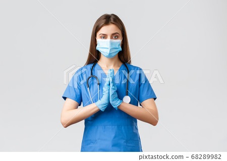 Covid-19, preventing virus, healthcare concept. Pray for medical workers. Female nurse or doctor in blue scrubs, medical mask, fighting coronavirus, press hands together hopeful, pleading Covid-19, preventing virus, healthcare concept. Pray for medical workers. Female nurse or doctor in blue scrubs, medical mask, fighting coronavirus, press hands together hopeful, pleading 68289982
