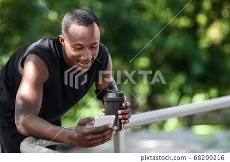 Smiling african sporty guy chatting on phone, drinking protein 68290216