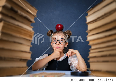 Happy smart girl in rounded glasses thoughtfully sitting between two piles of books with red apple on head, pointing finger and look at camera smiling Happy smart girl in rounded glasses thoughtfully sitting between two piles of books with red apple on head, pointing finger and look at camera smiling 68291565