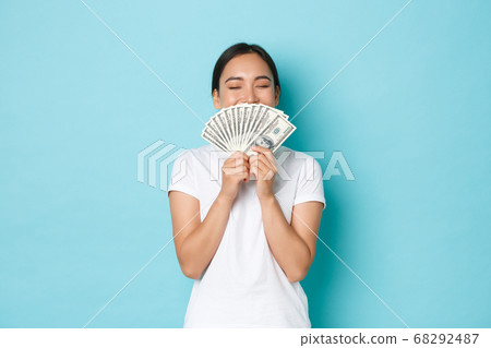 Shopping, money and finance concept. Young successful and lucky cheerful asian girl smelling dollars and smiling satisfied, enjoying being rich, holding cash over light-blue background 68292487