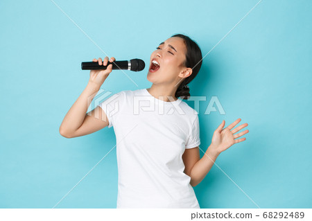 Lifestyle, people and leisure concept. Passionate and carefree pretty asian girl singing song in microphone, bending during performance, like going karaoke, standing light blue background performing Lifestyle, people and leisure concept. Passionate and carefree pretty asian girl singing song in microphone, bending during performance, like going karaoke, standing light blue background performing 68292489