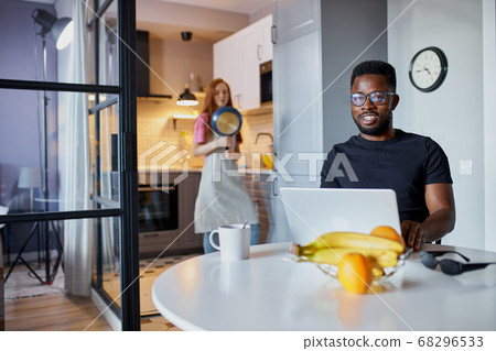 confident african man work on laptop at home while wife is busy in the kitchen confident african man work on laptop at home while wife is busy in the kitchen 68296533