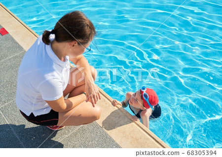 Woman trainer talking to girl child in swimsuit hat and goggles near outdoor pool 68305394