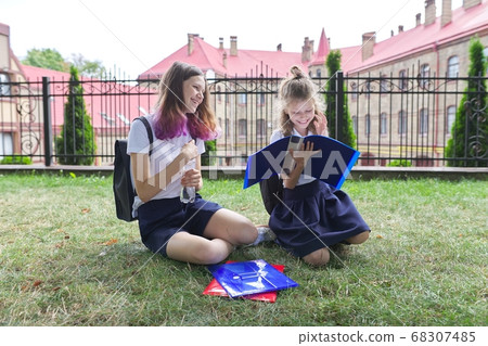 Two schoolgirls sitting on grass with books near school building, girls sisters Two schoolgirls sitting on grass with books near school building, girls sisters 68307485