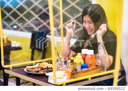 Asian woman sitting self isolate in steak restaurant and enjoy eating which have table shield plastic to separate another people from other customer when coronavirus outbreak,social distancing concept 68309322