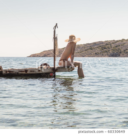 View of unrecognizable woman wearing big summer sun hat tanning topless and relaxing on old wooden pier in remote calm cove of Adriatic sea, Croatia 68330649