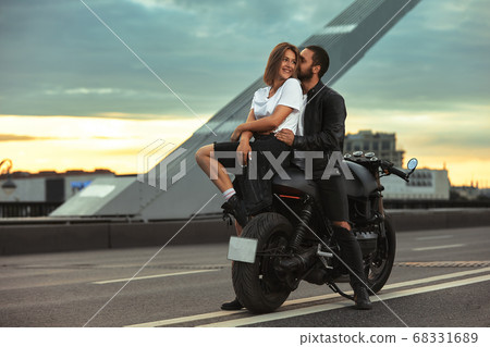 Young sexy woman hugging cute man in stylish black leather jacket, sitting on sports motorcycle on the bridge in the city on sunset and kissing Young sexy woman hugging cute man in stylish black leather jacket, sitting on sports motorcycle on the bridge in the city on sunset and kissing 68331689