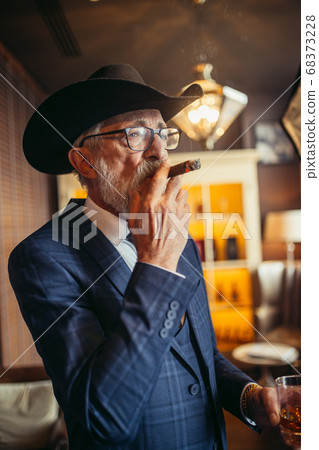 Portrait of old-aged male dandy in big hat smokes cigar and drinks alcohol. 68373228