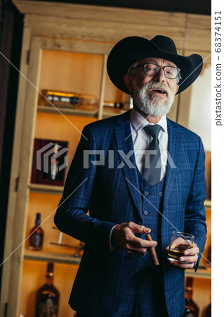 Portrait of old-aged male dandy in big hat smokes cigar and drin 68374151