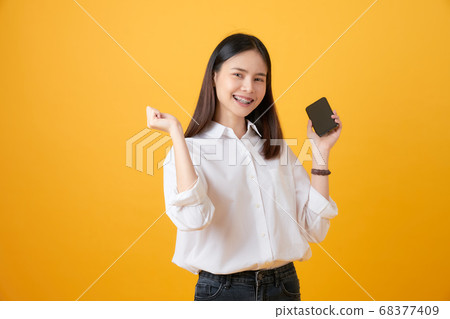 Cheerful beautiful Asian woman holding smartphone on light yellow background. 68377409