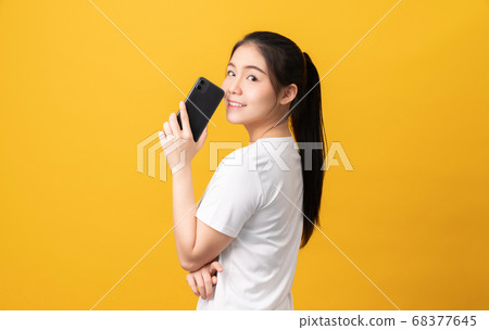 Cheerful beautiful Asian woman holding smartphone and typing message on light yellow background. Cheerful beautiful Asian woman holding smartphone and typing message on light yellow background. 68377645
