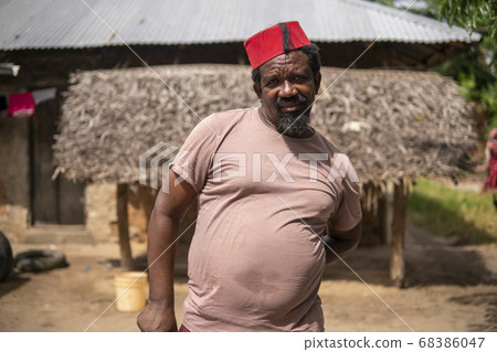 An African Older Man in Red Muslim Taqiyyah Fez Hat posing with a stick for lame people on Yard Near the Basic Hut with Thatched roof in Small Remote Village in Tanzania, Pemba island, Zanzibar 68386047