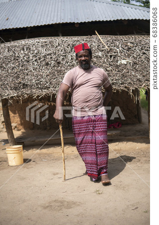 An African Older Man in Red Muslim Taqiyyah Fez Hat posing with a stick for lame people on Yard Near the Basic Hut with Thatched roof in Small Remote Village in Tanzania, Pemba island, Zanzibar 68386068