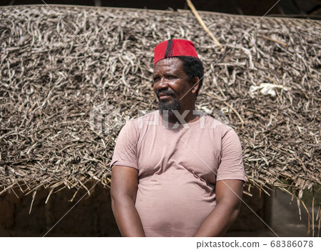 An African Older Man in Red Muslim Taqiyyah Fez Hat posing with a stick for lame people on Yard Near the Basic Hut with Thatched roof in Small Remote Village in Tanzania, Pemba island, Zanzibar 68386078
