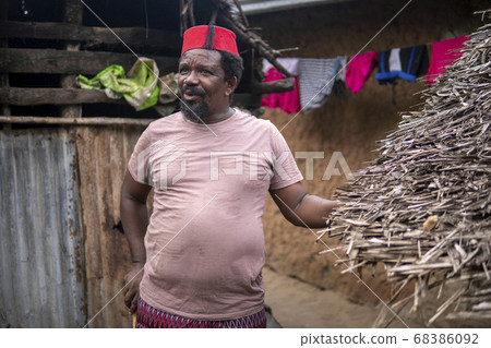 An African Older Man in Red Muslim Taqiyyah Fez Hat posing with a stick for lame people on Yard Near the Basic Hut with Thatched roof in Small Remote Village in Tanzania, Pemba island, Zanzibar 68386092