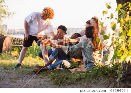 Group of friends clinking beer glasses during picnic at the beach. Lifestyle, friendship, having fun, weekend and resting concept. Group of friends clinking beer glasses during picnic at the beach. Lifestyle, friendship, having fun, weekend and resting concept. 68394934