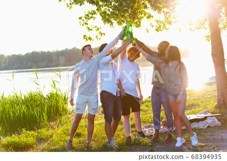 Group of friends clinking beer bottles during picnic at the beach. Lifestyle, friendship, having fun, weekend and resting concept. Group of friends clinking beer bottles during picnic at the beach. Lifestyle, friendship, having fun, weekend and resting concept. 68394935