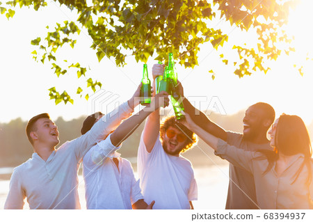 Group of friends clinking beer bottles during picnic at the beach. Lifestyle, friendship, having fun, weekend and resting concept. Group of friends clinking beer bottles during picnic at the beach. Lifestyle, friendship, having fun, weekend and resting concept. 68394937