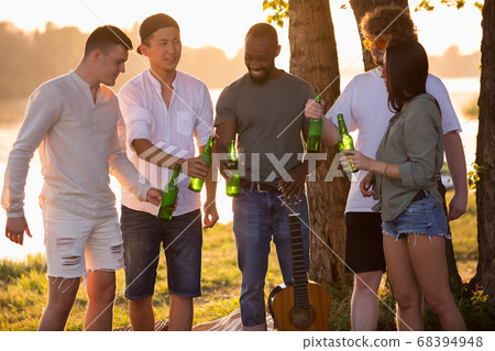 Group of friends clinking beer bottles during picnic at the beach. Lifestyle, friendship, having fun, weekend and resting concept. Group of friends clinking beer bottles during picnic at the beach. Lifestyle, friendship, having fun, weekend and resting concept. 68394948