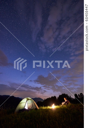 Lonely guy sitting by fire near tent on background of mountains under starry sky 68408447