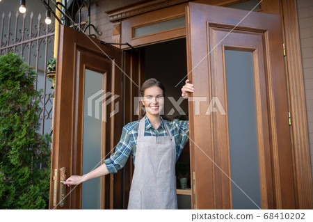 Smiling young waitress holding doors, welcoming clients Smiling young waitress holding doors, welcoming clients 68410202