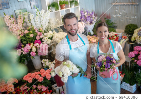 Bearded male and brown-haired female in aprons, holding bouquet and box of flowers, smiling 68410396