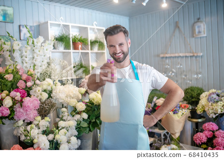 Smiling bearded male spraying water at camera inside flower shop 68410435