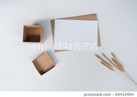Blank white paper on brown paper envelope with Bristly foxtail dry flower and Carton box. Mock-up of horizontal blank greeting card. Top view of Craft  envelope on white background. Flat lay minimal 68410994