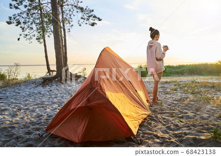 Charming young woman hiker drinking coffee on the beach Charming young woman hiker drinking coffee on the beach 68423138