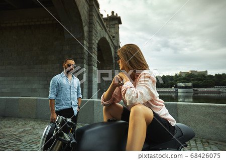 Romantic date on motorbike. Young woman sits on a motorcycle and looks at the man who comes to her. Couple in love with sunset under the bridge in the city Romantic date on motorbike. Young woman sits on a motorcycle and looks at the man who comes to her. Couple in love with sunset under the bridge in the city 68426075