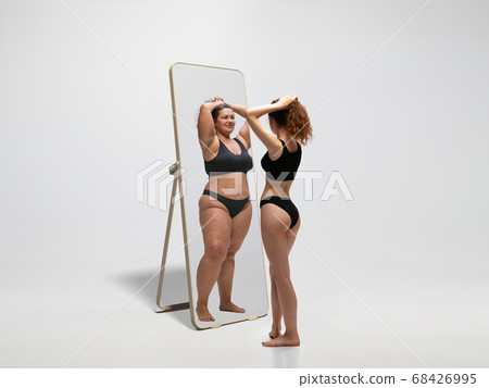 Young fit, slim woman looking at fat girl in mirror's reflection on white background Young fit, slim woman looking at fat girl in mirror's reflection on white background 68426995