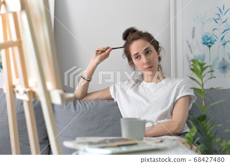 woman artist, dreams and ponders, thoughts in her head, at home on couch, a brush in her hand, a table with paints. Background, easel and wall with paintings. Looks at the painting. woman artist, dreams and ponders, thoughts in her head, at home on couch, a brush in her hand, a table with paints. Background, easel and wall with paintings. Looks at the painting. 68427480