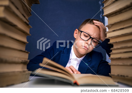 Smart boy in glasses sitting between two piles of books and read book thoughtfully 68428509