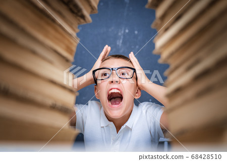 Young boy in stress getting crazy while studying and doing homework in children education concept sitting between two piles of books. 68428510
