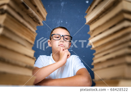 Smart boy in glasses sitting between two piles of books and look down thoughtfully Smart boy in glasses sitting between two piles of books and look down thoughtfully 68428522
