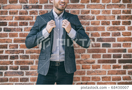 Suit man smart casual outfit young businessman adjusting collar of his blue wool blazer with shirt against brick wall background at office. Urban lifestyle 68430007