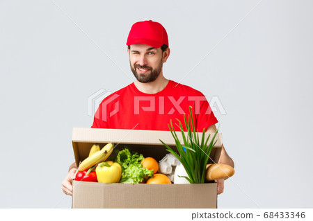 Groceries and packages delivery, covid-19, quarantine and shopping concept. Handsome smiling courier in red uniform, give cheeky wink as delivering food box, online order to client house 68433346