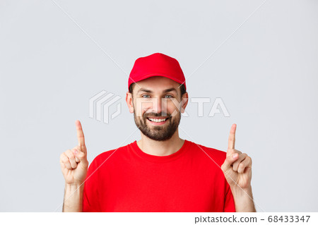 Online shopping, delivery during quarantine and takeaway concept. Cheerful bearded smiling courier in red uniform cap and t-shirt, invite take look at promo, pointing fingers up, grey background Online shopping, delivery during quarantine and takeaway concept. Cheerful bearded smiling courier in red uniform cap and t-shirt, invite take look at promo, pointing fingers up, grey background 68433347