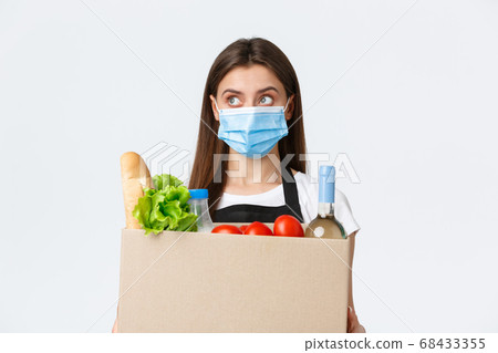Covid-19 social distancing, delivery and grocery shopping during coronavirus concept. Young pleasant saleswoman, cashier in medical mask handling over groceries to customer Covid-19 social distancing, delivery and grocery shopping during coronavirus concept. Young pleasant saleswoman, cashier in medical mask handling over groceries to customer 68433355