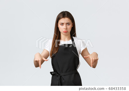 Small business, employees and coffee shop concept. Unhappy gloomy young saleswoman in black apron pointing fingers down and sulking disappointed. Displeased barista show bad news Small business, employees and coffee shop concept. Unhappy gloomy young saleswoman in black apron pointing fingers down and sulking disappointed. Displeased barista show bad news 68433358