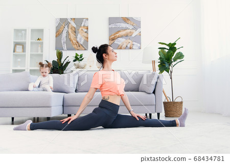 Happy calm mother doing morning exercises in yoga pose while her little daughter playing at home. Happy calm mother doing morning exercises in yoga pose while her little daughter playing at home. 68434781