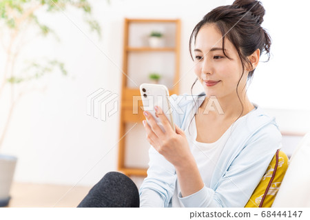 一個年輕的女人坐在沙發上,通過智能手機發送電子郵件 一個年輕的女人坐在沙發上,通過智能手機發送電子郵件 68444147