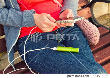 A girl uses a smartphone outdoors while charging A girl uses a smartphone outdoors while charging 68451396