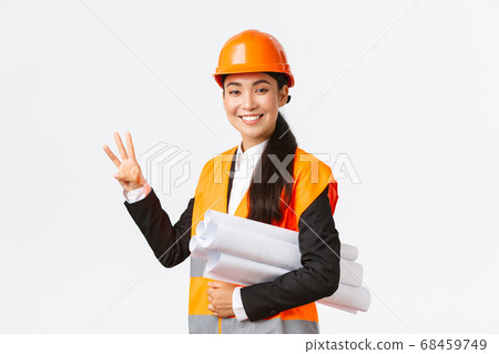 Smiling asian female architect at construction area, building estates, showing number three, carry blueprint as wearing safety helmet, giving fixed time for task done, white background Smiling asian female architect at construction area, building estates, showing number three, carry blueprint as wearing safety helmet, giving fixed time for task done, white background 68459749