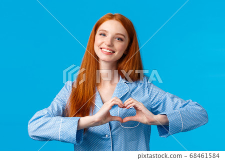 Relationship, love and tenderness concept. Cute sensual and feminine redhead girl long hair, wearing nightwear, showing heart gesture and smiling, showing affection or like, blue background 68461584